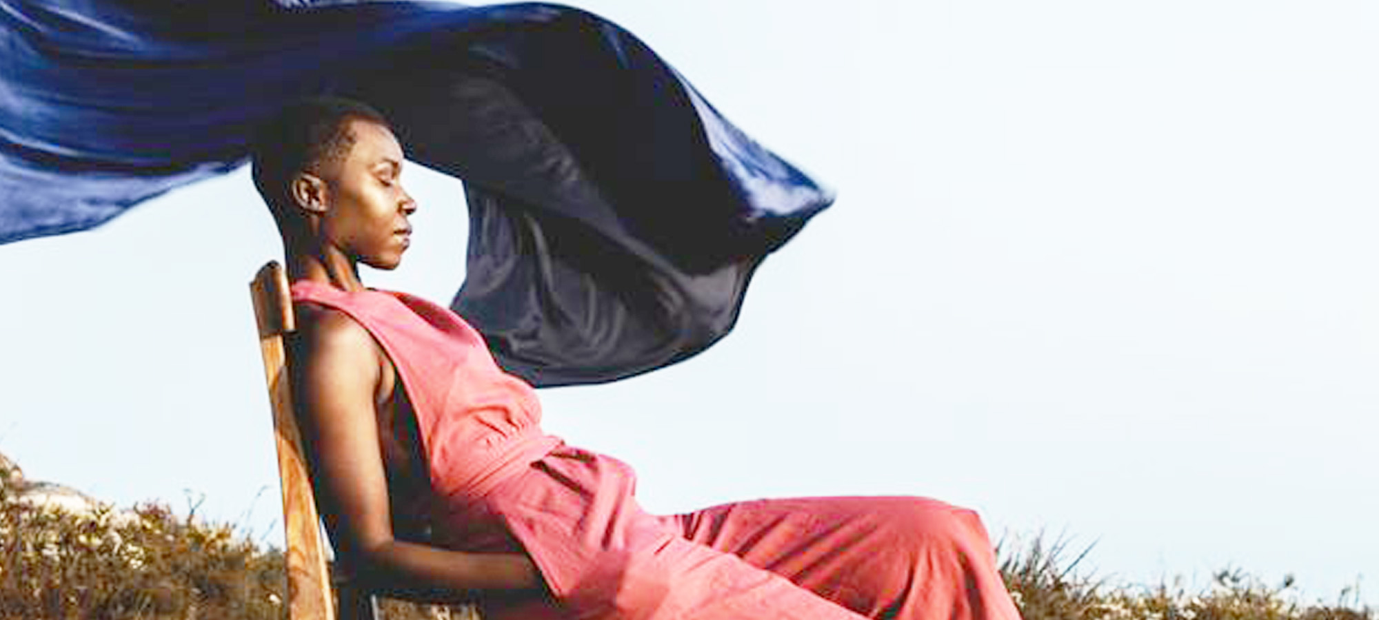 A woman in a red jumpsuit closes her eyes while sitting on a chair. A navy scarf blows in the background.