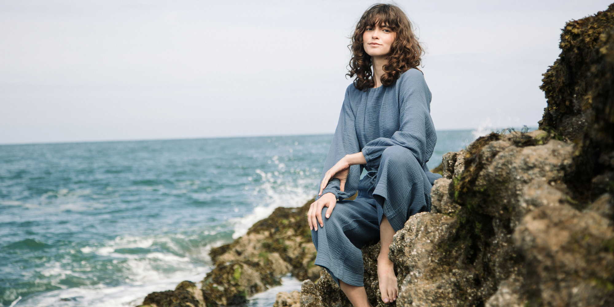 Someone perched on rocks by the sea wearing a blue jumpsuit by Beaumont Organic.