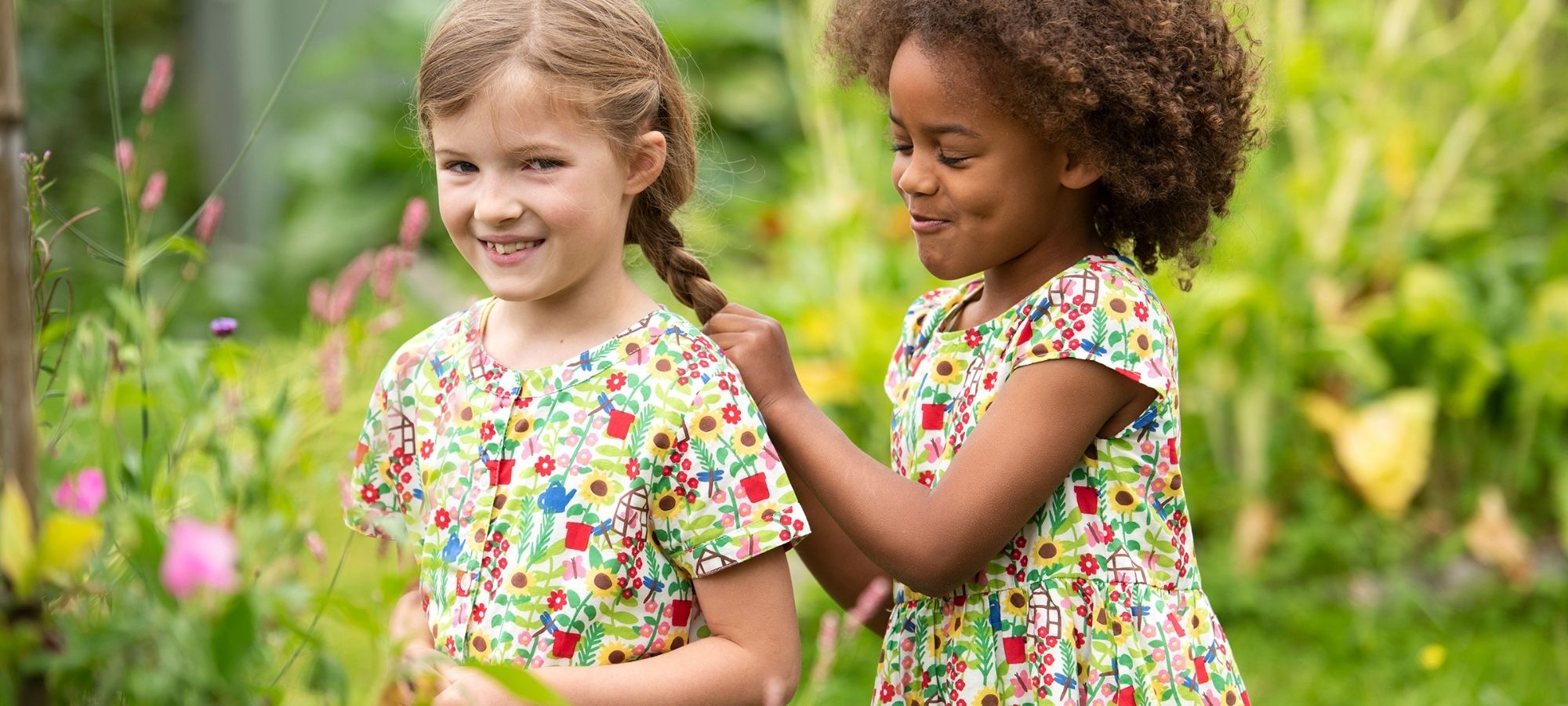 Two children in a garden wearing bright floral ethically made dresses by Frugi.