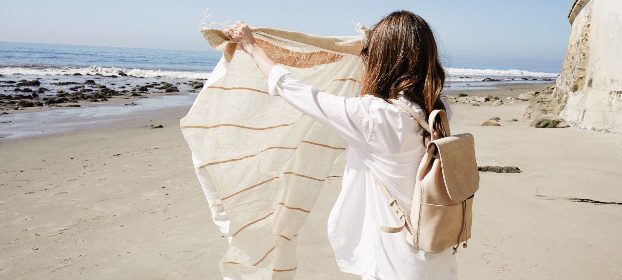 Someone unfolding a striped cotton towel on a beach, wearing a light leather backpack by Parker Clay.