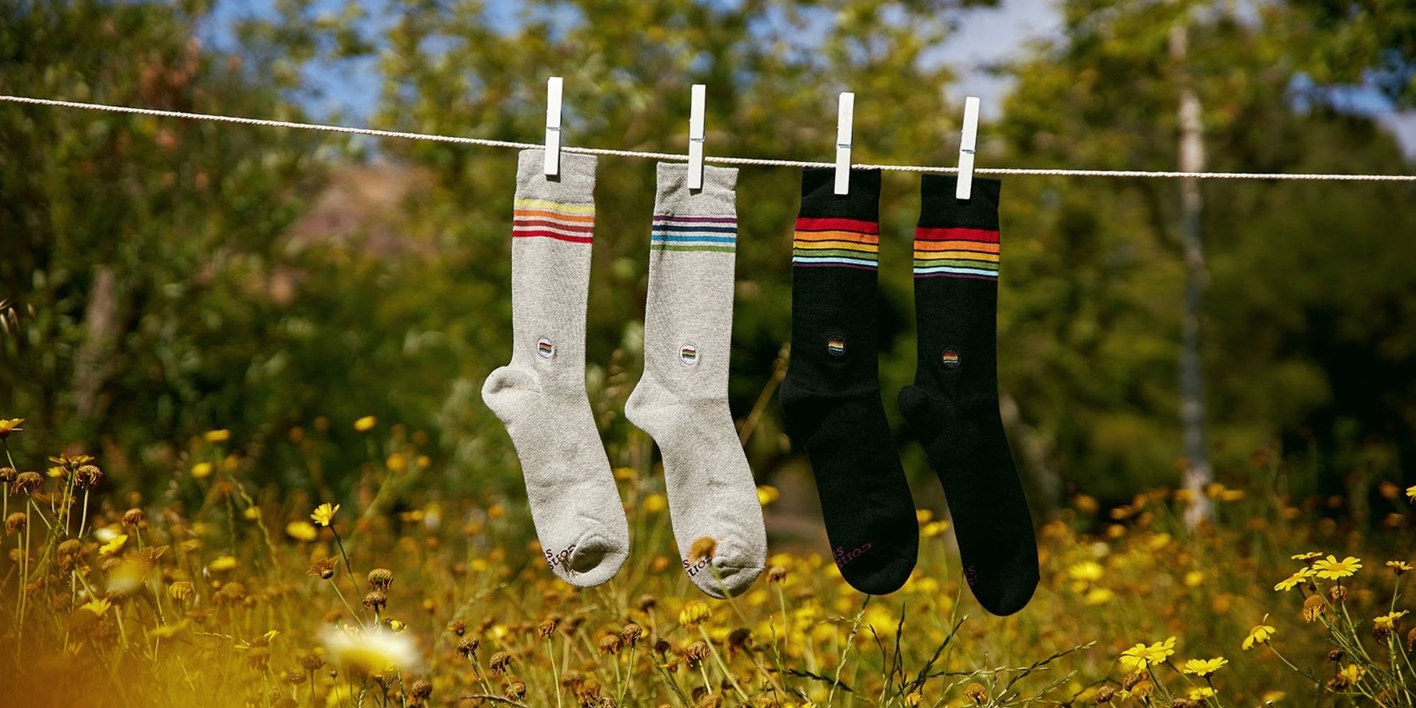 Two pairs of socks hanging on a line outside with rainbow graphics, one grey and one black. Ethically made by Conscious Step.