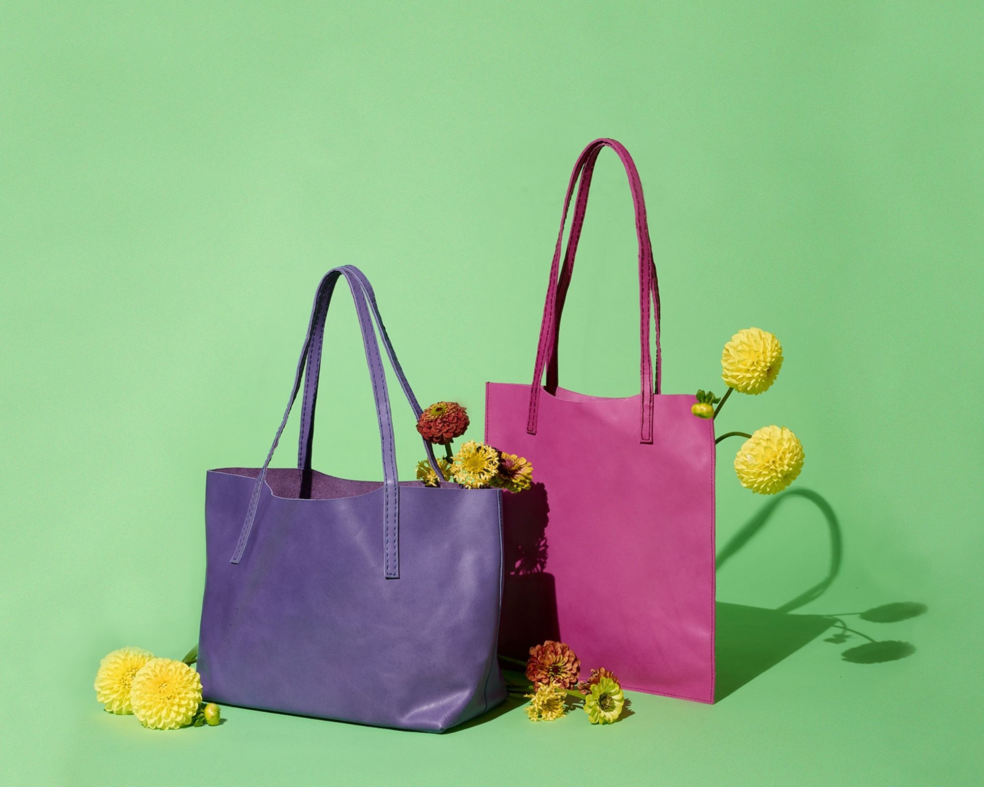 A purple and pink leather handbag by Raven + Lily filled with flowers against a green backdrop.