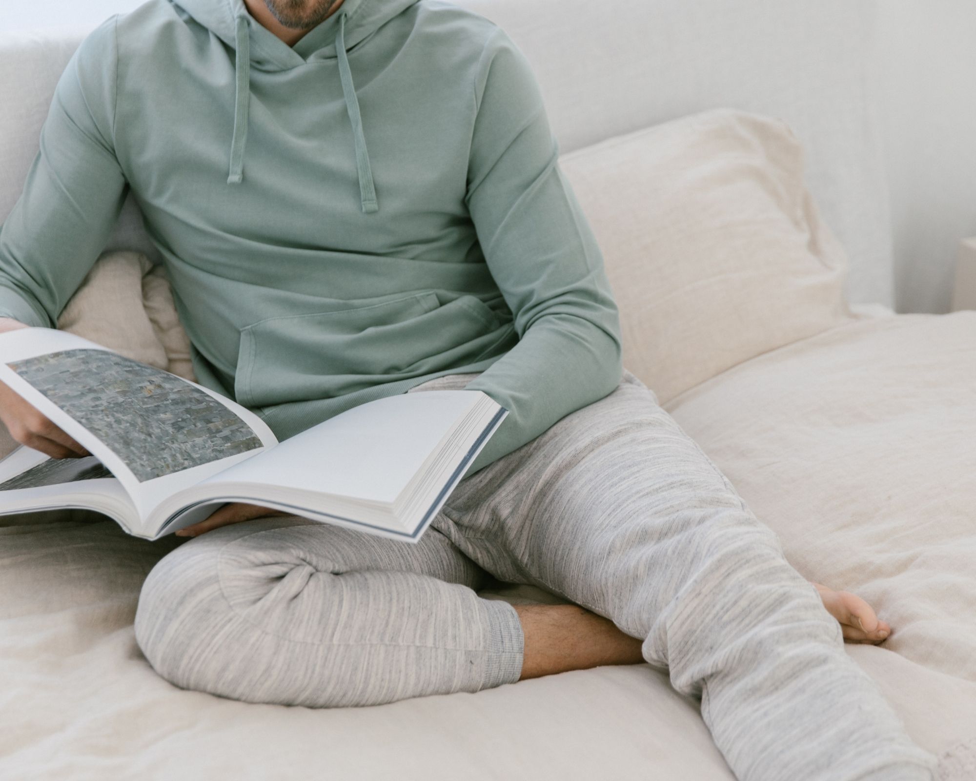 A cropped shot of someone sitting on a bed wearing a sage green eco-friendly hoodie and light grey sweatpants ethically made by tentree.