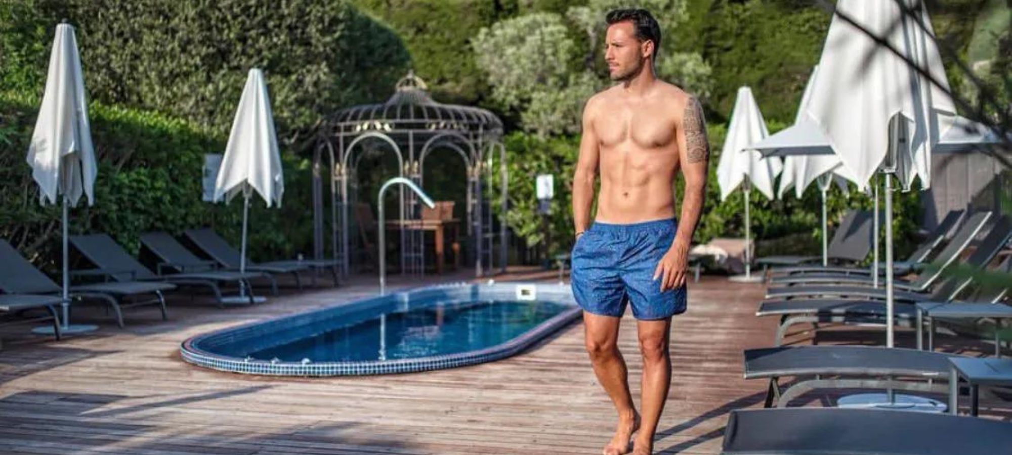 Someone wearing men's swimwear standing by a pool by True Tribe.