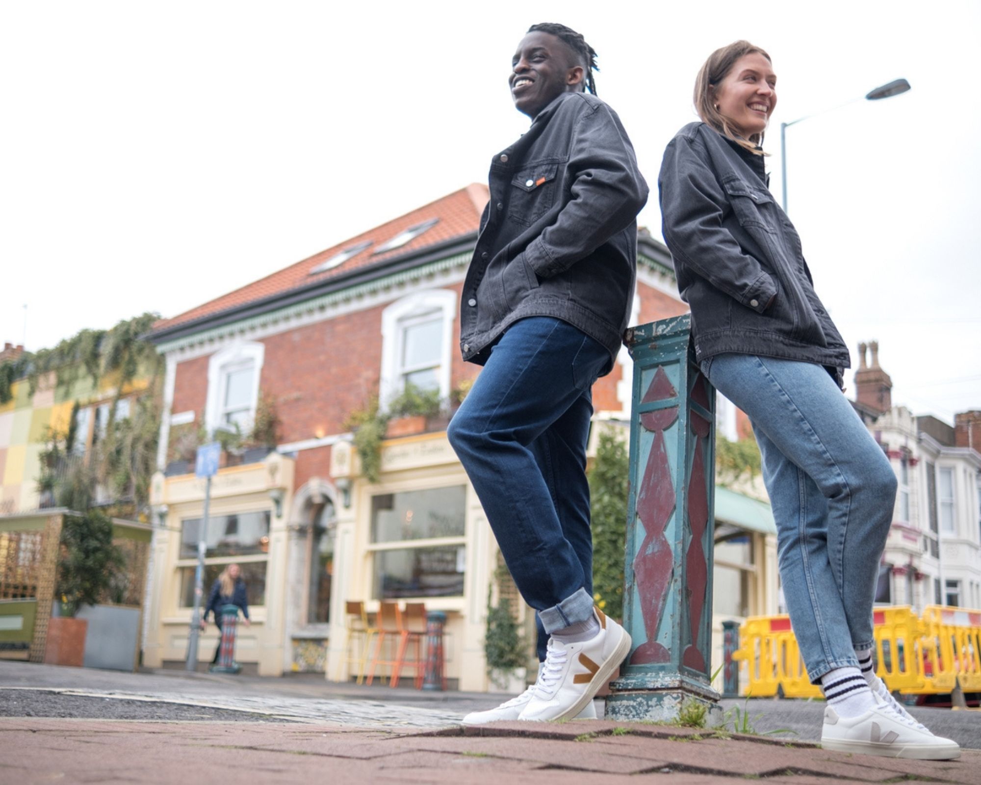 Two people smiling and leaning on a pole outside wearing ethical and sustainable denim jackets and jeans by Yes Friends.