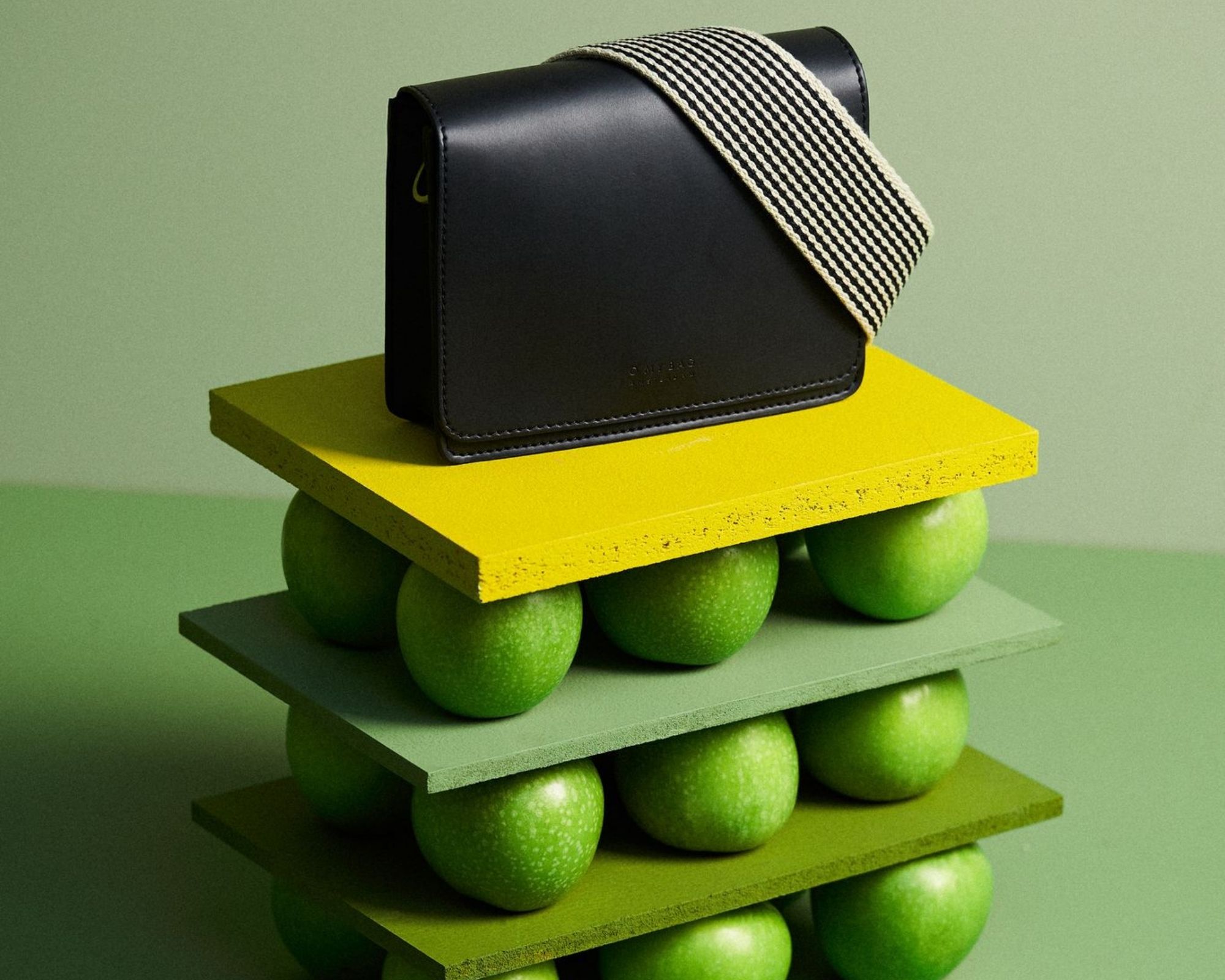 A small black O My Bag handbag made of apple leather sitting on boards stacked with green apples.