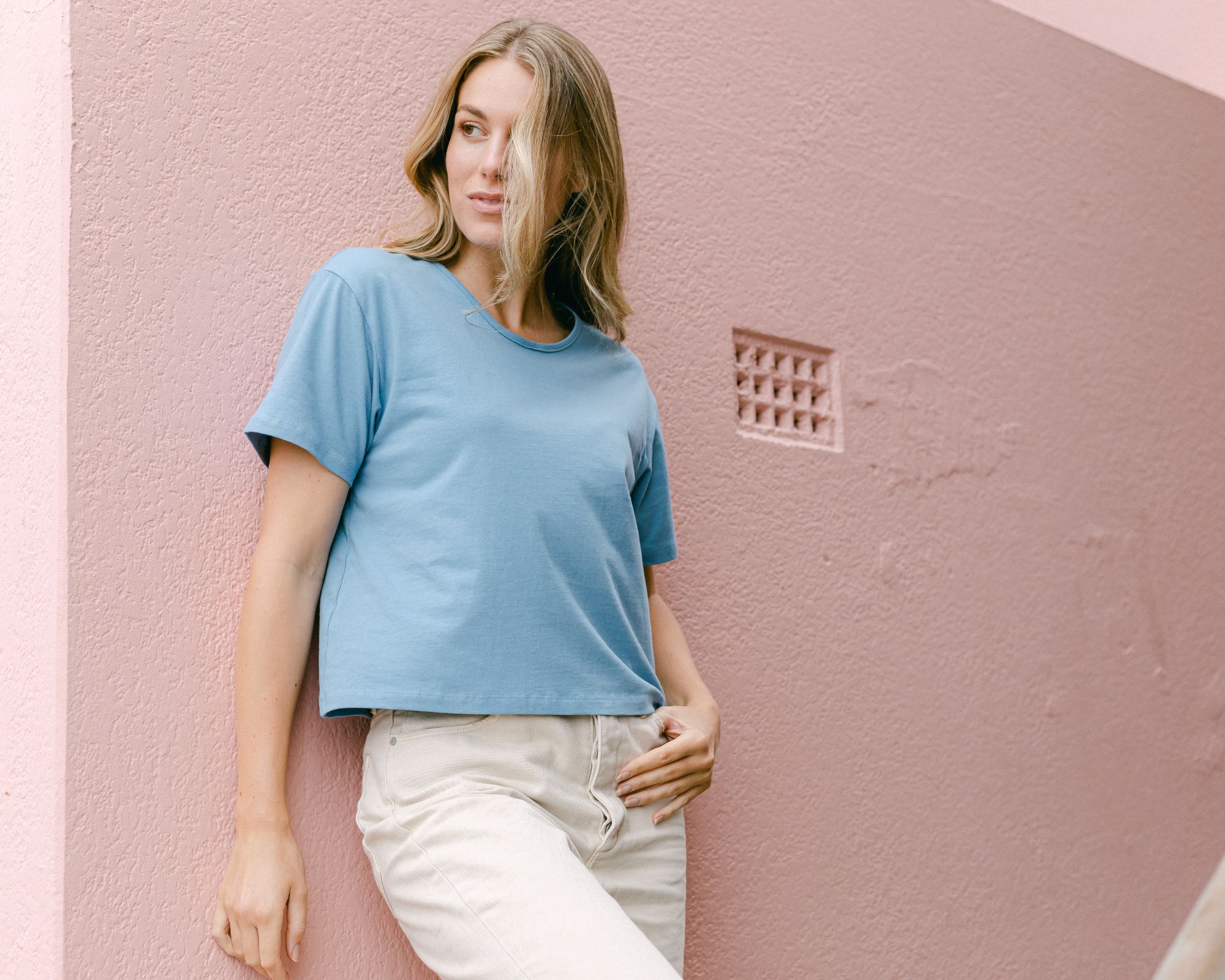Person wearing a light blue custom fit crop t-shirt by Citizen Wolf, leaning against a pink wall.