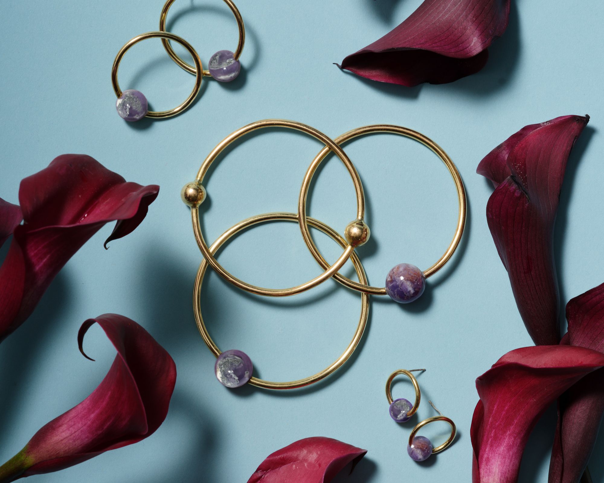Ethically made jewellery laying near red petals on a blue background, by Raven + Lily.