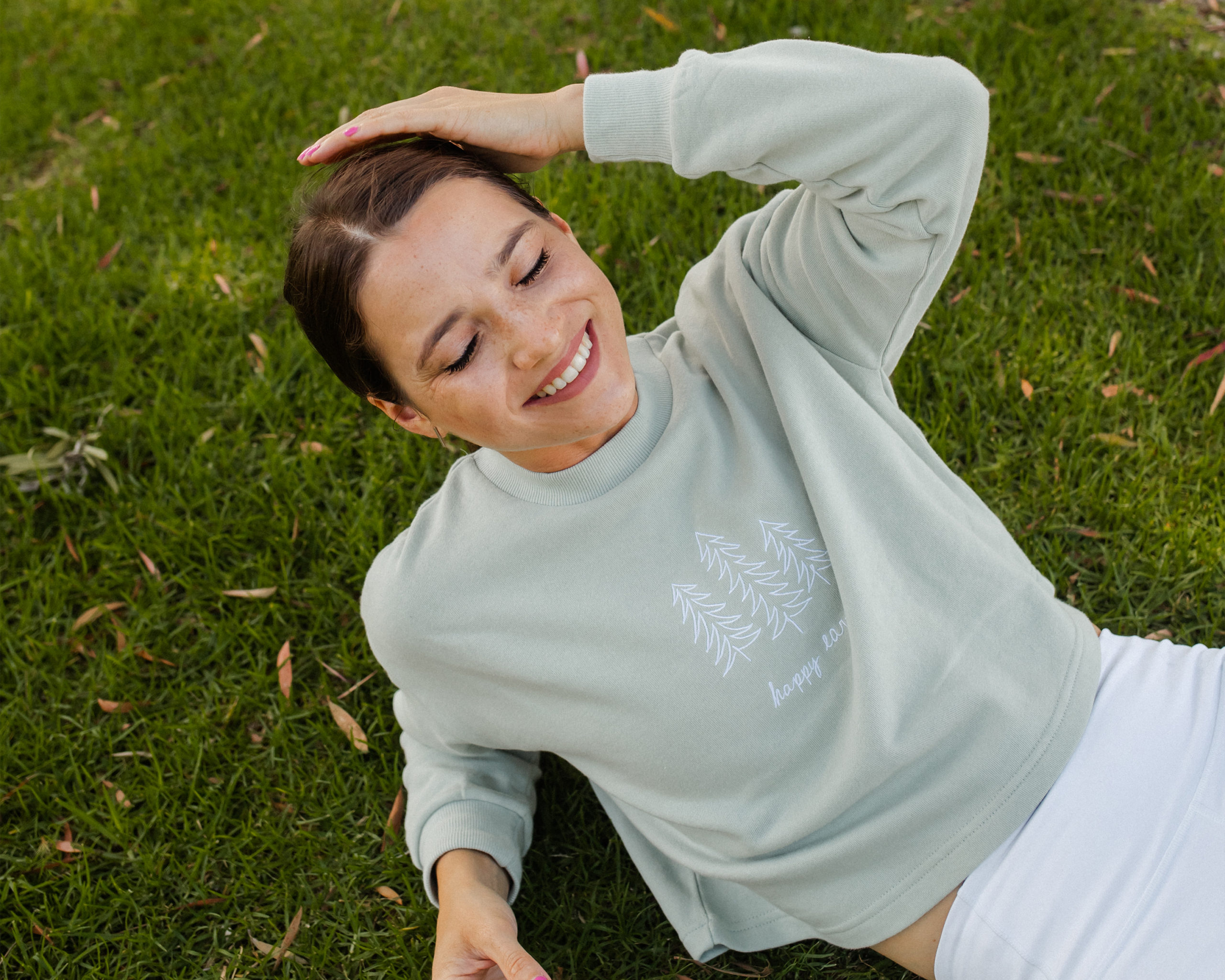 Someone looking happy reclined on grass wearing a pale green Happy Earth sweater.