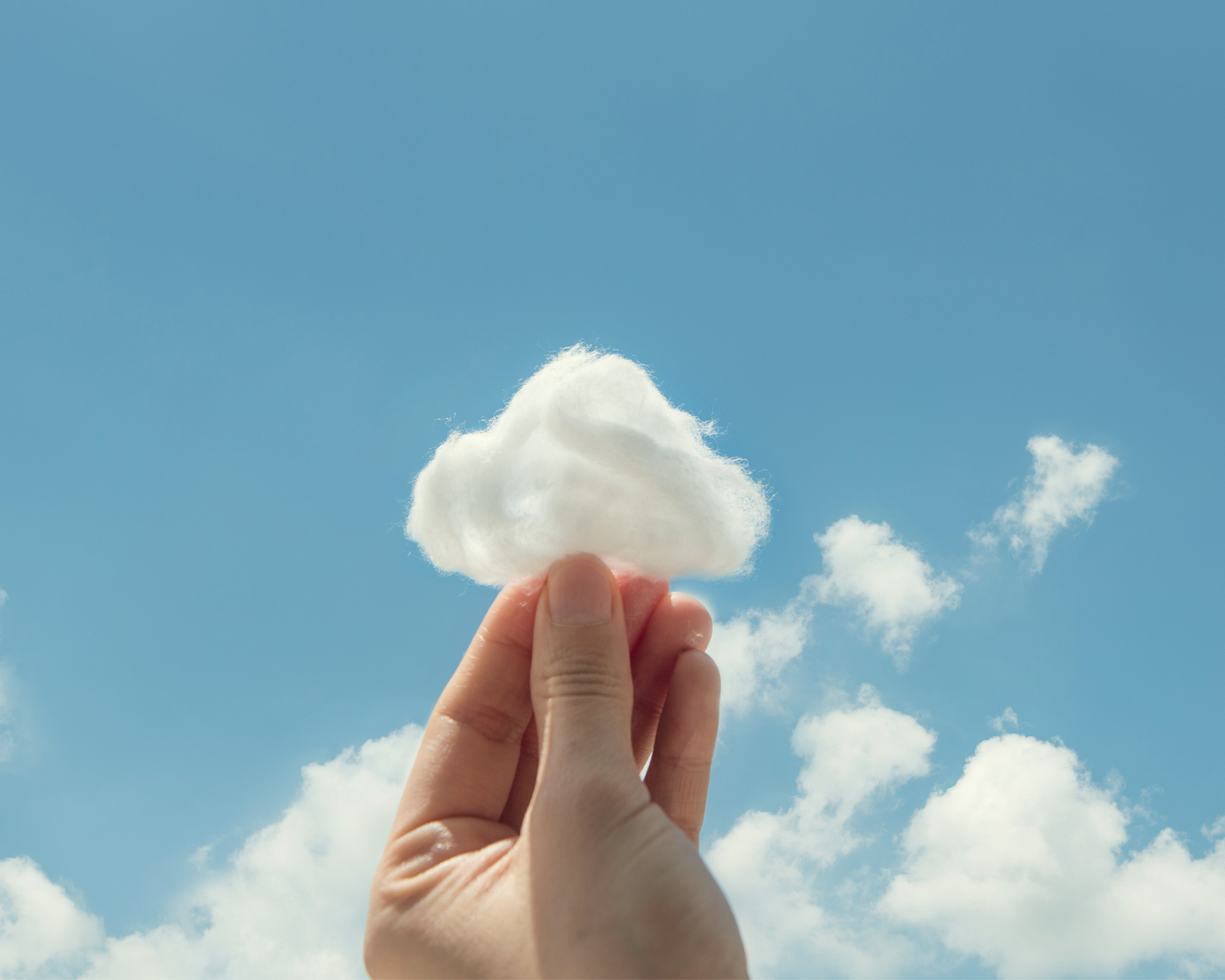 Hand holding a bit of cotton against a cloudy blue sky, the cotton looking like an extension of the clouds.