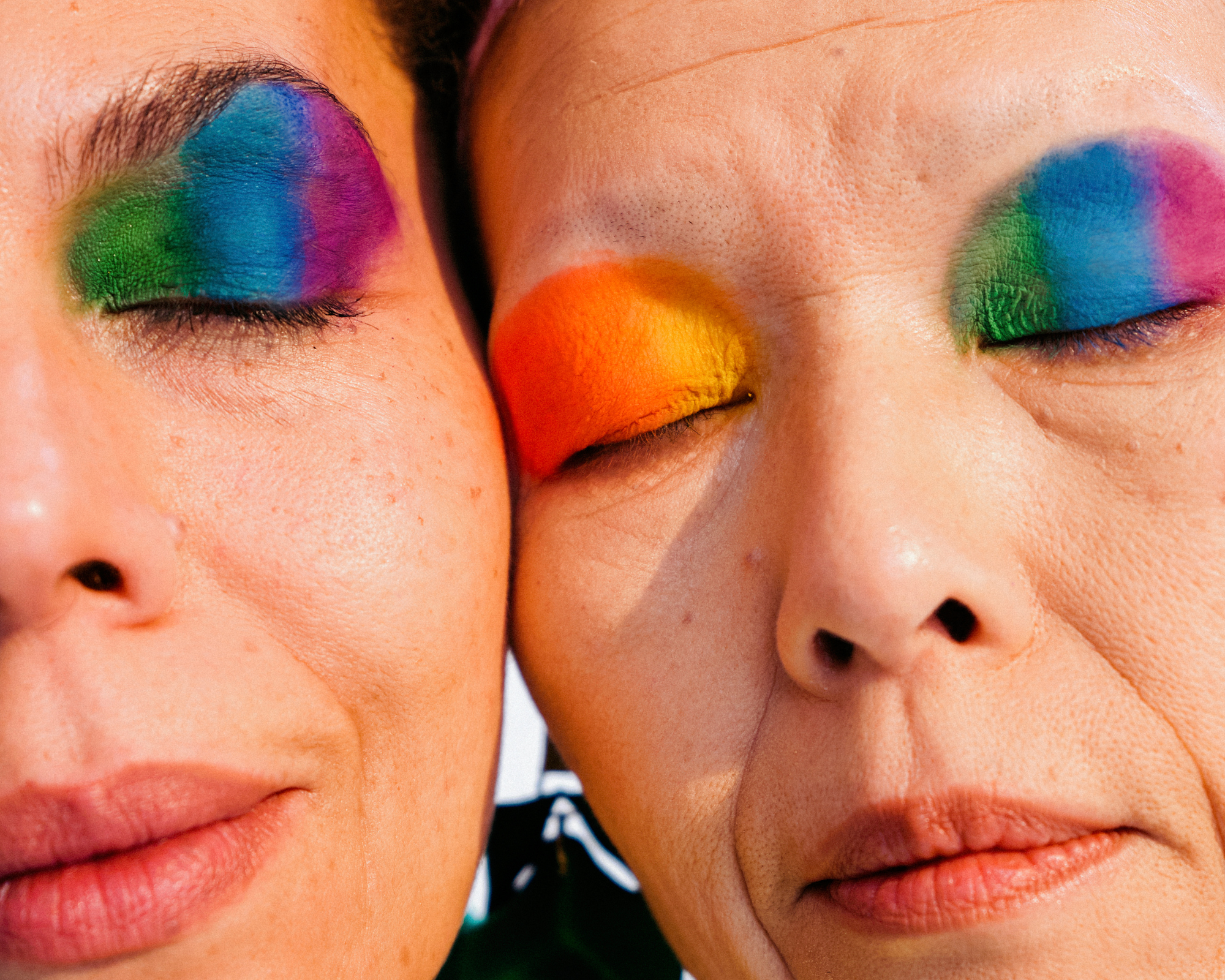 Close-up photo of two people with eyes closed, wearing vibrant rainbow eyeshadow in blue, purple, green, and orange tones.